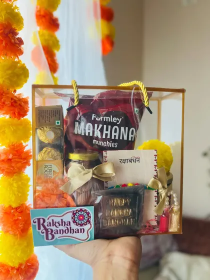 This clear box hamper includes a custom "Happy Raksha Bandhan" chocolate bar, adding a sweet personal touch.