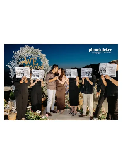 A creative group shot where friends and family hold up newspapers to announce the couple's engagement, with the couple sharing a kiss in the center.