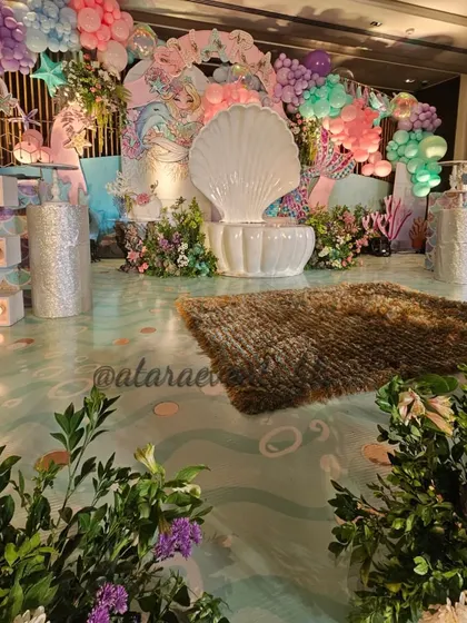 A magical mermaid-themed first birthday stage. The setup features a large white seashell seat, pastel balloon garlands, and an underwater-inspired floor, creating a dreamy scene for the birthday girl.
