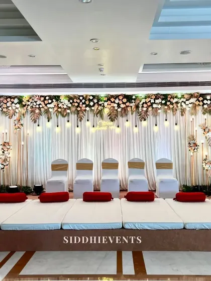An all-white wedding stage featuring traditional floor seating, a canopy of white and blush florals, and hanging light bulbs for a serene and elegant atmosphere.