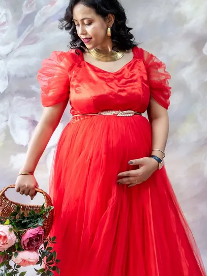 A lovely studio portrait against a floral painted backdrop. The mom-to-be looks elegant in a red tulle gown, holding a basket of fresh flowers.