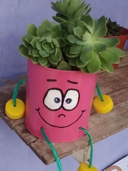 This cute pink planter with a worried expression sits on a wooden shelf, holding a healthy succulent. It’s a simple, charming piece made from an upcycled can.