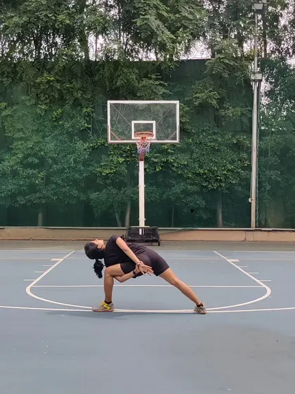 Holding Baddha Parsvakonasana (bound side angle pose) on a basketball court surrounded by greenery. This deep stretch for the hips and shoulders feels even better with fresh air and open space.