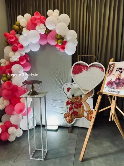 A lovely naming ceremony setup featuring a pink and white balloon arch. The arrangement includes a teddy bear cutout, a pedestal for a cake or ceremony items, and an easel displaying a photo of the baby.