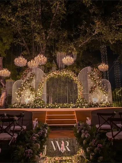 This stunning engagement stage was set under a canopy of trees, with chandeliers hanging like stars. The centerpiece is a modern, white slatted structure, with a floral arch creating a focal point for the couple's ceremony.
