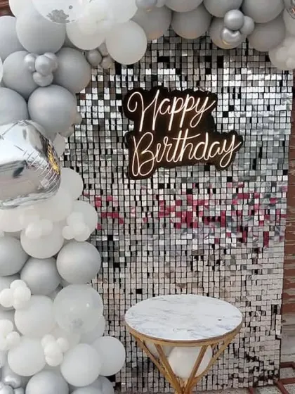 A birthday setup with a silver shimmer wall, a 'Happy Birthday' sign, and a balloon garland in silver and white.