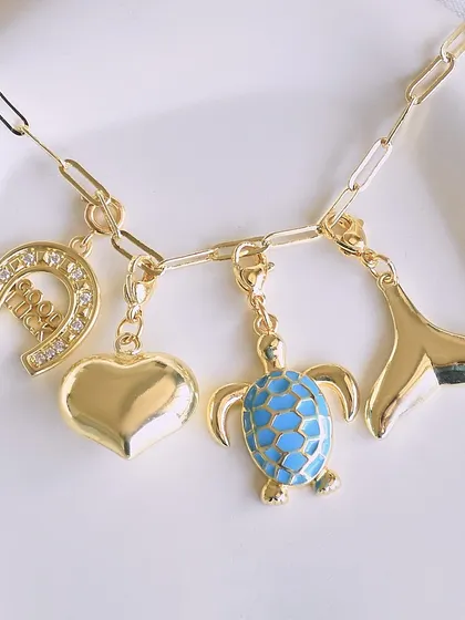 Under the sea vibes with this charm collection. It features a good luck horseshoe, a puffy heart, a blue enamel turtle, and a whale tail charm on a gold link chain.
