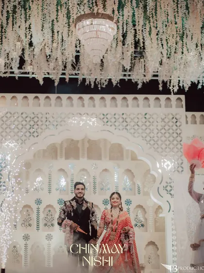 The couple's grand entrance against a stunning white backdrop with intricate jaali work and a floral ceiling. Cold pyro effects add to the magical moment.
