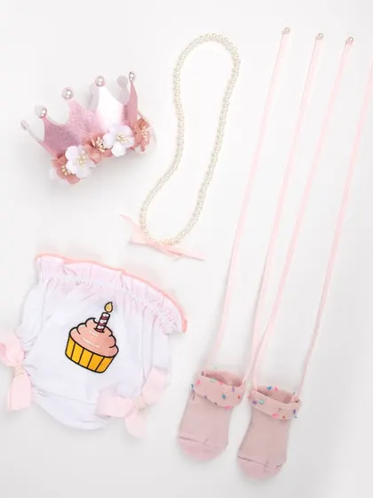 A baby's first birthday outfit set, including a cupcake-themed bottom, pink socks, a pearl necklace, and a floral crown.