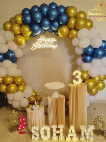 A clean and modern birthday setup with a blue, gold, and white balloon ring, corrugated plinths, and a neon sign.