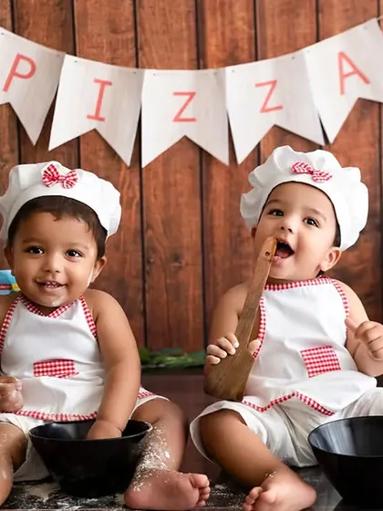 A fun twin photoshoot with a "Pizza Kitchen" theme, where two babies are dressed as little chefs.