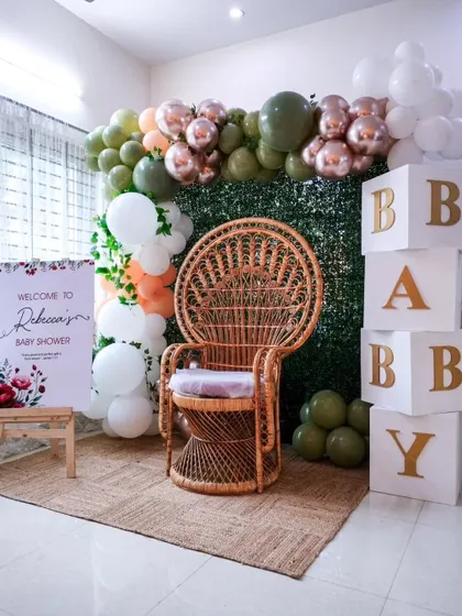 An elegant at-home baby shower. This setup features a green hedge wall, a peacock chair for the mom-to-be, and "BABY" blocks, transforming a simple corner into a beautiful photo spot.