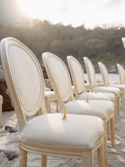 Simple elegance for a ceremony by the Ganges. Classic white chairs are lined up on the sand, each with a small golden bell for guests to ring, creating a serene and beautiful aisle.