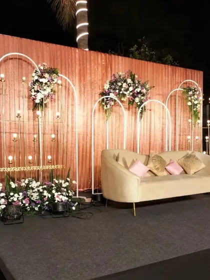We designed this sophisticated reception stage with a rose-gold sequin backdrop, framed by modern white arches and accented with floral bouquets. The curved lounge seating adds a contemporary and comfortable touch for the couple.