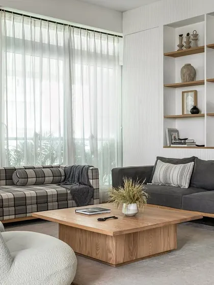 A modern farmhouse aesthetic defines this living room. The space blends natural elements like a fluted wall with built-in oak shelves, a comfortable grey sofa, and low-height white boucle chairs to encourage intimate conversations.