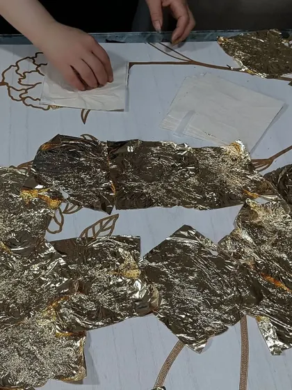 The intricate process of hand-applying genuine metallic leaf to our 'Flourish' wood veneer wallcovering. This image captures the moment of creation, where skilled artisans transform a surface into a luxurious work of art.