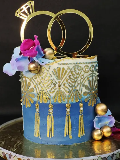 An engagement cake with a unique, bohemian-inspired design. I used a tassel-like stencil effect with gold detailing on a blue ombre buttercream base, topped with a modern ring-shaped acrylic topper.