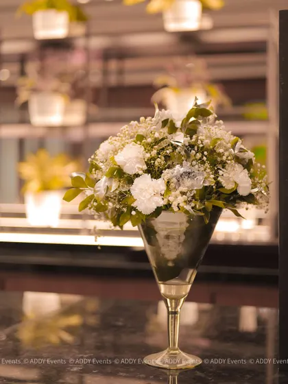 An elegant floral arrangement in a silver martini glass vase. The bouquet of white carnations and baby's breath is a classic choice for sophisticated events like anniversary dinners or formal receptions.