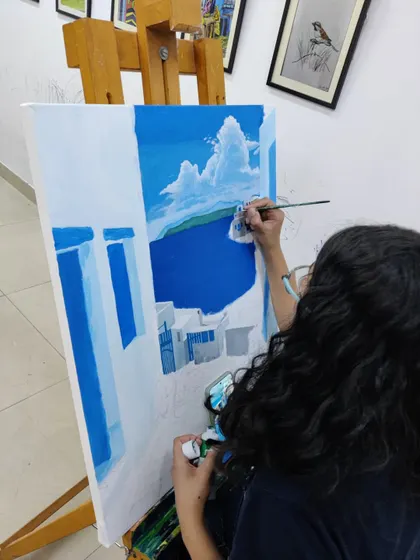 The student adds clouds to the sky, using white paint to create a sense of depth and atmosphere in her acrylic on canvas piece.