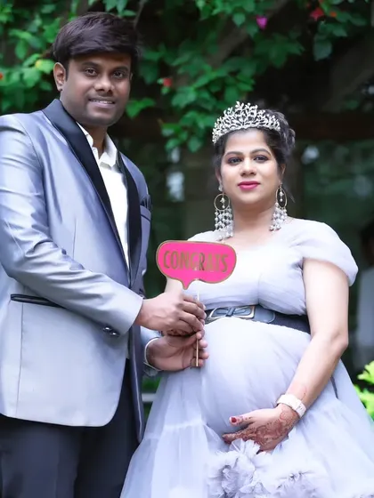 A sweet couple's maternity photo, with the father-to-be holding a "Congrats" sign, both dressed in stylish grey and silver outfits.