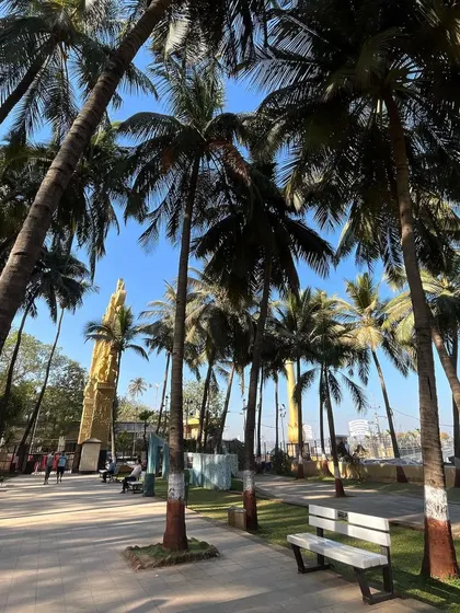 A wide shot of the walkway at Shivaji Park, lined with tall palm trees and leading towards the memorial pillar.