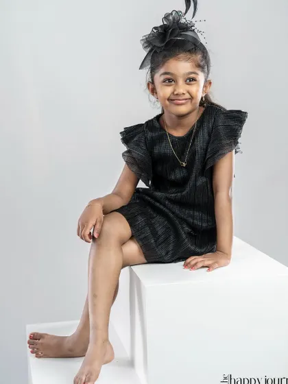 A stylish young girl in a black dress and fascinator, posing with confidence on a white block in the studio.
