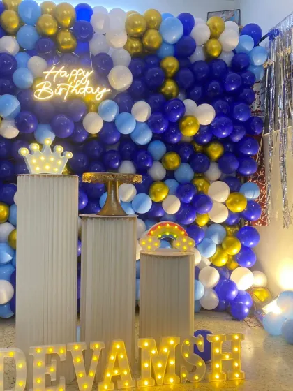 A birthday setup with a full wall of blue, white, and gold balloons. The decor includes a neon sign, white plinths, and marquee letters.
