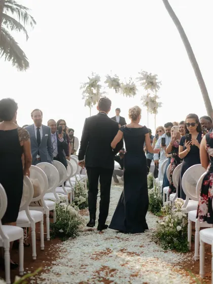 A view from behind as the groom and his mother walk down the aisle, showing the guests and the beautiful floral arch awaiting them.