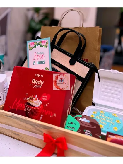 A lovely birthday gift set in a wooden tray, featuring Plum BodyLovin' products, a mini "bento box" cake, a stylish handbag, and a heartfelt greeting card.