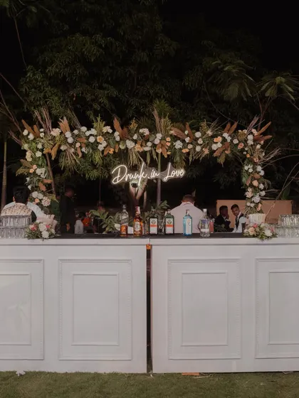 A chic and stylish bar setup for an outdoor Sangeet, featuring a 'Drunk in Love' neon sign and a floral arch with pampas grass.