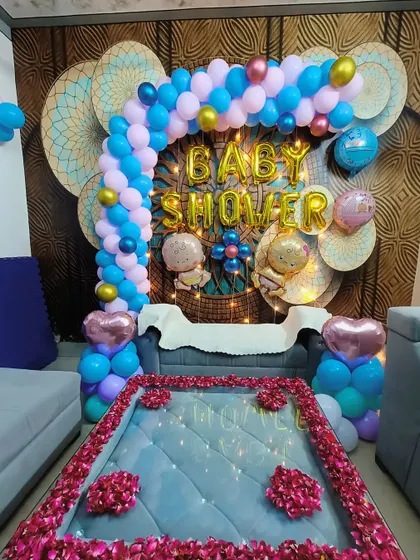 A lovely baby shower setup in a home setting. The decor features a blue and pink balloon arch over the sofa, with rose petals on the table, creating a cozy yet celebratory atmosphere.
