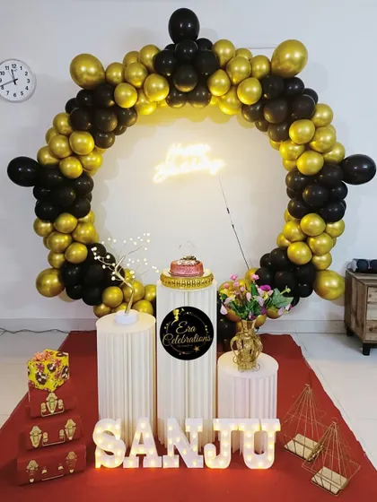 An elegant home birthday decoration with a circular balloon arch in black and gold. A neon 'Happy Birthday' sign and light-up name letters add a personalized, warm glow.