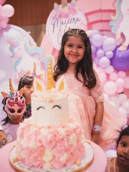 The birthday girl, Maseera, smiling brightly next to her adorable unicorn-themed cake.