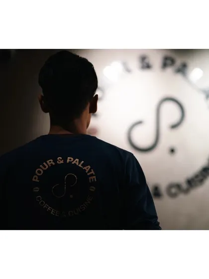 A shot from behind a barista at Pour & Palate cafe, with the cafe's logo softly focused in the background. It's a subtle branding shot that captures the atmosphere of the place.