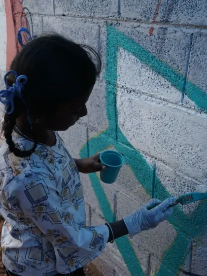 A young girl with blue ribbons in her hair meticulously paints a section of the wall. These projects are as much about building confidence and learning new skills as they are about creating a beautiful mural.