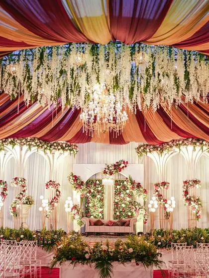 A grand reception stage with a ceiling of red and gold drapes, a massive floral chandelier, and elegant white structures.