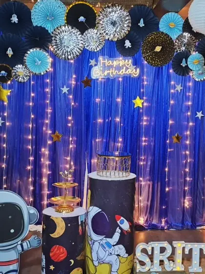 An eco-friendly space theme for Srithik's first birthday. This creative setup uses paper fans and a fabric backdrop with fairy lights to create a starry night sky, complete with an astronaut cutout and themed pedestals.