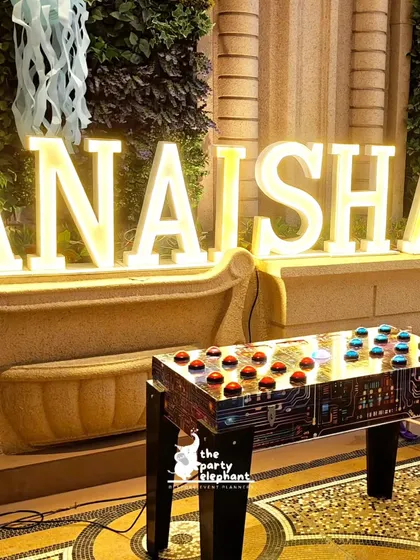 Large, illuminated marquee letters spelling out "ANAISHA" in front of a bench, paired with an interactive game table for guests to enjoy.