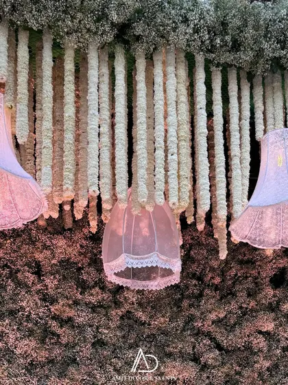 A close-up of the hanging decor, showing delicate pink lampshades interspersed with strings of white baby's breath against a textured floral wall.