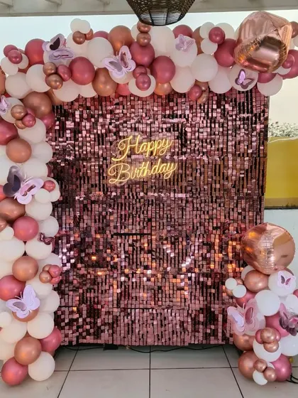 A beautiful rose gold sequin wall backdrop for a terrace party. The setup is framed with a white and rose gold balloon arch, butterfly accents, and a 'Happy Birthday' neon sign.