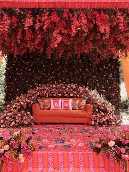 A breathtaking Mehendi stage drenched in shades of red and orange. The setup features a dense canopy of flowers and a beautifully detailed floral carpet.