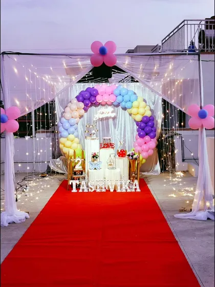 A magical terrace decoration featuring a red carpet entrance and a canopy of sheer curtains with fairy lights. The rainbow balloon arch adds a splash of color.