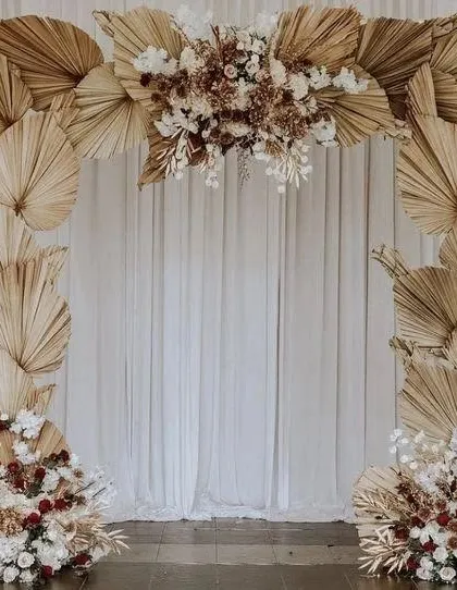 An elegant and simple boho backdrop idea. The arch is crafted entirely from dried palm leaves and accented with white and neutral florals, creating a beautiful, textured frame for a ceremony or photo corner.
