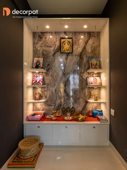This pooja niche uses a dark marble-finish back panel to create a dramatic effect. The white floating shelves and base cabinet provide a clean contrast and functional storage.