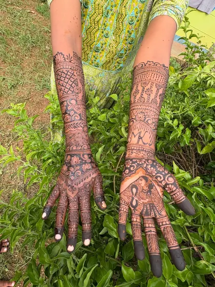 A beautiful bridal mehendi with personalized elements. One palm features a dulha-dulhan portrait, while the other has the couple's initials 'MK'. The arms are decorated with traditional motifs, creating a complete story.