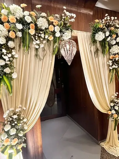 An elegant doorway treatment with an arch of white and peach flowers, soft cream-colored drapes, and a hanging crystal lantern. This classic design creates a graceful and welcoming entrance.