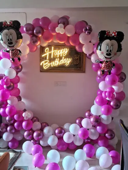 A Minnie Mouse themed birthday party decoration. I used a circular balloon arch with pink, purple, and white balloons, Minnie Mouse foil balloons, and a central 'Happy Birthday' neon sign.