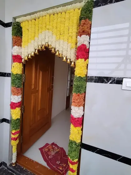 A vibrant main door decoration using thick garlands of colorful flowers. The arch is adorned with small brass bells that jingle as guests arrive.