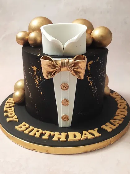 We present the essence of formal sophistication with our black and gold tuxedo cake. This design features a sharp, edible shirt collar and a lustrous gold bow tie set against a matte black tier, accented with gold splatters and spheres, making it an impeccable choice for a gentleman's milestone birthday.