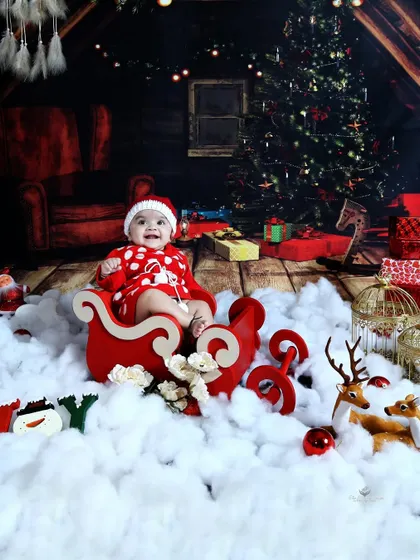 A joyful baby in a sleigh, surrounded by 'snow' and presents. The rustic, warm-toned background creates a cozy, cabin-like Christmas atmosphere.